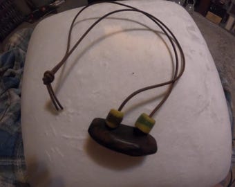 May include: A brown leather necklace with a dark wooden pendant. The pendant has two yellow and green beads attached. The necklace is knotted at the top and is laying on a white surface.