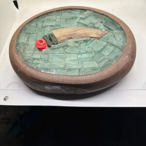 May include: A round, handmade ceramic dish with a mosaic design. The interior features a mosaic of light green glass pieces, with a wooden element and a red rose. The rim is a dark brown color.