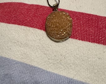 May include: A pendant necklace featuring a large, oval-shaped, amber-colored stone with a textured surface. The stone is set in a silver-colored metal frame and hangs from a dark brown cord. The background is a striped fabric in red, white, and blue.