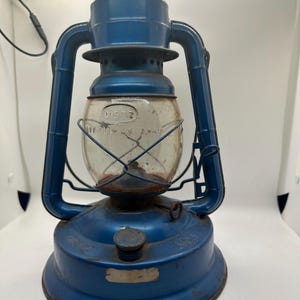 May include: A vintage blue Dietz No. 10 lantern with a clear glass globe. The lantern has a metal frame and handle, and the word "DIETZ" is visible on the glass. The lantern is designed to be used with oil.