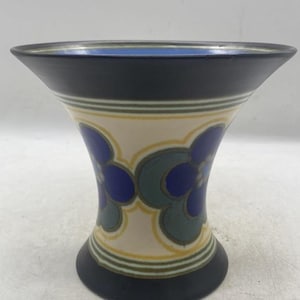 May include: A decorative ceramic vase with a flared top and a wide base. The vase features a black rim and base, with a cream-colored body adorned with blue and green floral patterns. Yellow and green stripes encircle the body.