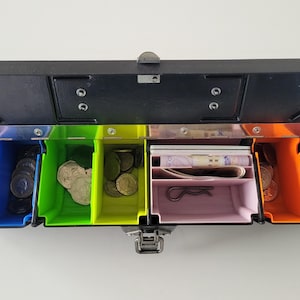 May include: A black cash register with open, rainbow-coloured compartments. The compartments contain coins, banknotes, and a bobby pin. The cash register is designed for organising money.