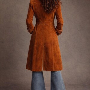 May include: A long, rust-colored suede coat with a tailored fit, featuring a button-down front and a flared skirt. The coat is paired with blue denim bell-bottom jeans. The model's back is facing the camera.