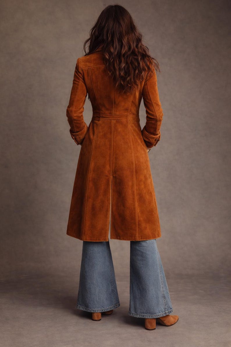 May include: A long, rust-colored suede coat with a tailored fit, featuring a button-down front and a flared skirt. The coat is paired with blue denim bell-bottom jeans. The model's back is facing the camera.