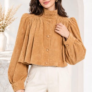 Op de afbeelding: Een beige blouse met suède-look, voorzien van een hoge kraag, geplooide schouders en volumineuze lange mouwen met knoopmanchetten. Het kledingstuk heeft een kort, uitlopend silhouet met verticale plooien vanaf het schouderstuk.