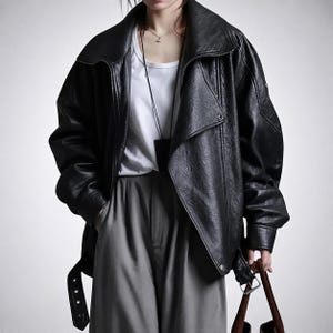Peut inclure: Veste en cuir noire avec un large col et une taille ceinturée. La veste est portée sur un haut blanc et un pantalon gris. Un sac en cuir marron est tenu à la main.