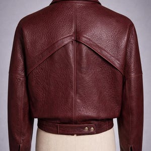 May include: A burgundy leather jacket displayed on a mannequin. The jacket features a textured finish, a collar, and a unique design element on the upper back. It has a belt detail at the waist with two buttons and buttoned cuffs.