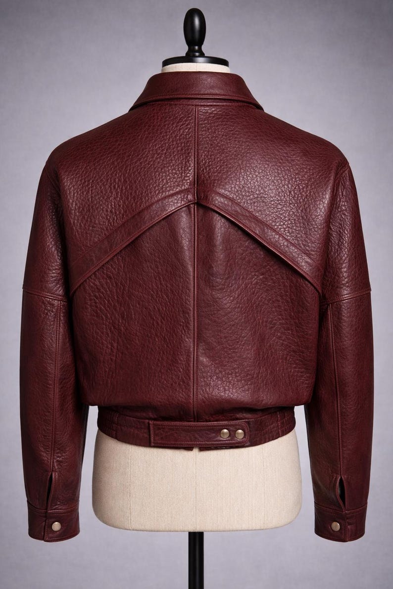 May include: A burgundy leather jacket displayed on a mannequin. The jacket features a textured finish, a collar, and a unique design element on the upper back. It has a belt detail at the waist with two buttons and buttoned cuffs.