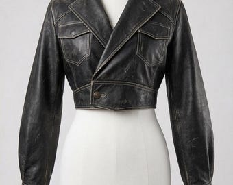 May include: A cropped black leather jacket featuring a distressed finish, a wide notched collar, and long sleeves with cuffs. The jacket includes two chest pockets with flaps and a single button closure at the front waist, showcasing visible stitching.
