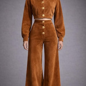 May include: A two-piece corduroy outfit in a warm brown hue. The set includes a cropped jacket with gold buttons and wide-leg pants, creating a retro-inspired look. The jacket has long sleeves and a round neckline.
