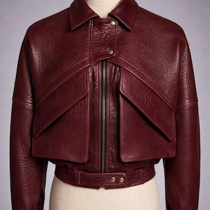 Puede incluir: Chaqueta de cuero burdeos con cuello, dos bolsillos delanteros y cierre de cremallera. La chaqueta tiene un diseño corto con detalle de cinturón en la cintura y se muestra en un maniquí.