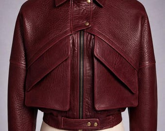 May include: A burgundy leather jacket with a collared neckline, two front pockets, and a zip-up closure. The jacket has a cropped design with a belt detail at the waist and is displayed on a mannequin.
