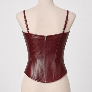 May include: Burgundy-colored corset with adjustable straps and a full-length zipper closure down the back. The bodice features vertical seams for shaping. The corset is displayed on a mannequin.