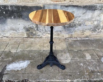Puede incluir: Una mesa de bistró redonda con un tablero circular construido con tablones de madera en tonos marrón claro y oscuro alternados. La mesa se apoya en una base de pedestal de metal negro, delgada y con un pie decorativo de tres puntas.