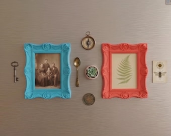 May include: A collection of decorative magnets and framed art pieces on a stainless steel refrigerator. Includes a blue frame with a family portrait, a red frame with a fern print, and a vintage key.