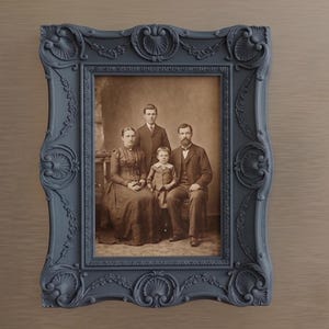 May include: Two framed prints with ornate, dark blue frames. One features a sepia-toned family portrait, the other a botanical print of a fern. The frames have detailed scrollwork and are mounted on a neutral-toned wall.
