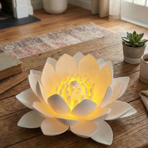 Puede incluir: Una lámpara blanca con forma de flor de loto, con un brillo amarillo cálido que emana del centro. La lámpara está sobre una mesa de madera, con una planta suculenta en maceta y una taza de té cerca. Los pétalos de la lámpara están en capas.