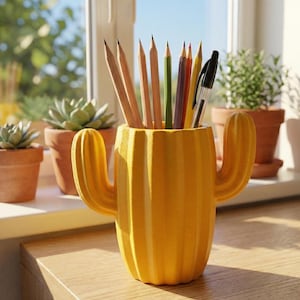 May include: A yellow cactus-shaped desk organizer holding pencils and a pen. The organizer is made of a ribbed material and has two arms. The pencils are various colors, and the pen is black. The organizer sits on a wooden surface.
