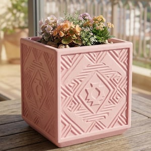 Puede incluir: Una maceta cuadrada de color rosa claro con un patrón geométrico, llena de flores coloridas y pequeñas piedras. La maceta está hecha de un material sólido y se encuentra sobre una superficie de madera.
