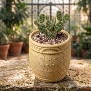 May include: A light yellow, patterned plant pot containing a cactus with green, paddle-shaped leaves. The pot is filled with soil and small stones. The background shows a greenhouse with other potted plants, creating a natural setting.
