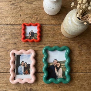 May include: Three wavy-edged picture frames in red, light pink, and dark green, each containing a photograph of a couple. The frames have a smooth, rounded, and undulating border design. The red frame is square, while the pink and green frames are rectangular.