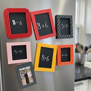 Puede incluir: Una colección de siete marcos de fotos magnéticos rectangulares y cuadrados en rojo, negro, rosa, amarillo y naranja. Cinco marcos tienen centros negros con texto blanco que indica los tamaños 8x6, 5x7, 5x5 y 4x6 cm. Un marco contiene una foto.