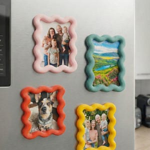 Könnte beinhalten: Vier rechteckige magnetische Bilderrahmen mit gewellten, geschwungenen Rändern in Pastellrosa, gedecktem Türkis, leuchtendem Orange und Sonnengelb. Jeder Rahmen enthält ein anderes Foto, darunter Familienporträts und eine Landschaft.