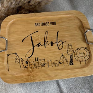 Könnte beinhalten: Eine rechteckige Bambus-Brotdose mit hellbraunem Finish. Der Deckel trägt den Namen "Jakob" in Schreibschrift, darüber der Text "BROTDOSE VON". Darunter sind Cartoon-Tiere eingraviert. Metallverschlüsse sichern den Deckel.