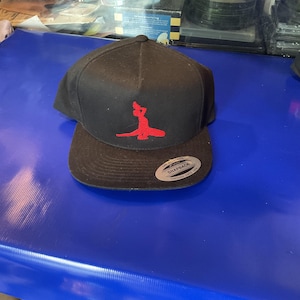 May include: Black snapback hat with a red graphic on the front. The hat has a flat brim and a snap closure at the back. The hat is sitting on a blue surface.