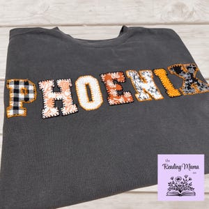 May include: A dark grey sweatshirt with the name "PHOENIX" appliquéd across the front. The letters are made of various patterned fabrics, including plaid, floral, and paisley designs. The sweatshirt is folded on a light wood surface.