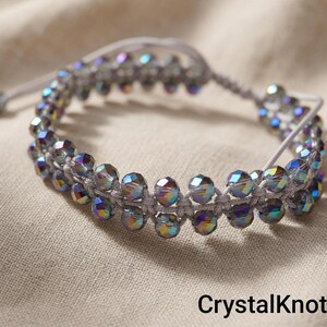 Puede incluir: Una pulsera hecha a mano con cuentas de cristal iridiscentes tejidas sobre un cordón gris. La pulsera ajustable tiene un diseño de macramé y se muestra sobre una tela de tono neutro. El texto "CrystalKnotBG" es visible en la parte inferior.