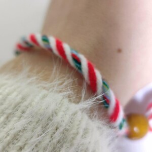 Puede incluir: Una pulsera trenzada con hilos rojos, blancos y verdes. La pulsera se lleva en la muñeca, con una cuenta amarilla. El texto "CrystalKnotBG" está en la parte inferior de la imagen.