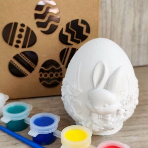 Può includere: Un uovo di Pasqua in ceramica bianca con un design a coniglietto, accompagnato da vasetti di vernice nei colori verde acqua, blu, giallo e rosso, e un pennello. Sullo sfondo, una scatola marrone con ritagli a forma di uovo.