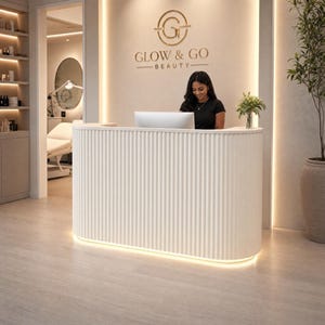 May include: A white, curved reception desk with a vertical fluted texture and integrated base lighting. The desk is positioned in front of a wall featuring the gold-colored text GLOW & GO BEAUTY and a circular logo above a computer monitor.