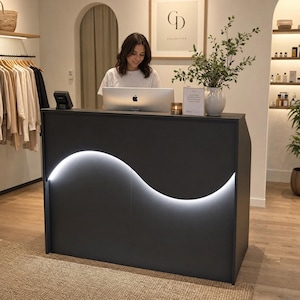 May include: A dark gray reception desk featuring a prominent, curved, illuminated white light strip across the front panel. The desk is positioned in a retail space with a computer, a small candle, and a potted green plant on the top surface.
