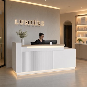 May include: A white reception desk with a vertical ribbed front panel and a light wood countertop. The wall behind features the backlit text LUMIÈRE AESTHETIC MEDICINE. A vase with greenery sits on the desk next to a computer monitor.
