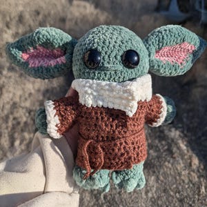 K&ouml;nnte beinhalten: Handgefertigte H&auml;kelpuppe einer Figur mit gro&szlig;en schwarzen Augen, gro&szlig;en Ohren und einem braunen Mantel mit wei&szlig;em Besatz. Die Puppe hat einen gr&uuml;nen Kopf und gr&uuml;ne F&uuml;&szlig;e sowie einen wei&szlig;en Kragen. Die Puppe ist etwa 15 cm gro&szlig;.