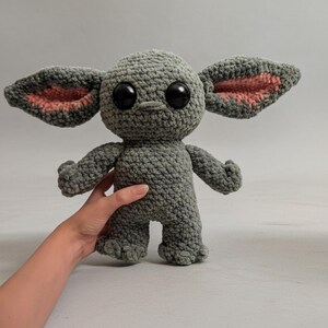 K&ouml;nnte beinhalten: Eine graue H&auml;kelpuppe mit gro&szlig;en schwarzen Augen und &uuml;bergro&szlig;en Ohren mit rosa Innendetails. Die Puppe hat kleine Arme und F&uuml;&szlig;e. Die Puppe wird von der Hand einer Person gehalten.
