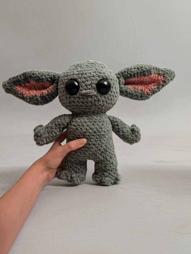 K&ouml;nnte beinhalten: Eine graue H&auml;kelpuppe mit gro&szlig;en schwarzen Augen und &uuml;bergro&szlig;en Ohren mit rosa Innendetails. Die Puppe hat kleine Arme und F&uuml;&szlig;e. Die Puppe wird von der Hand einer Person gehalten.