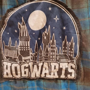 May include: A blue and black plaid shirt with a patch featuring a silhouette of Hogwarts castle against a starry night sky. The patch has the word "HOGWARTS" in white lettering.