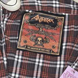 May include: A brown, red, and white plaid flannel shirt with a large patch featuring the band name "Anthrax" and the phrase "The Greater of Two Evils." The patch has a skull and lightning bolt design. A pair of gray sneakers is visible.