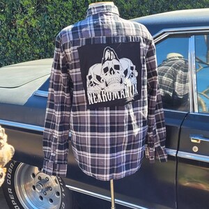 May include: A black and white plaid flannel shirt with a black patch on the back. The patch features a skull design with the text "NEKRUMANTIX".