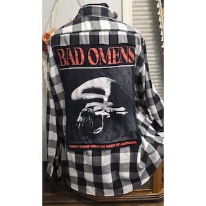 May include: A black and white buffalo plaid long-sleeve button-down shirt featuring a large rectangular patch on the back. The patch displays the text "BAD OMENS" in red, a black and white graphic of a figure, and the text "I DON'T KNOW WHAT I'M MADE OF ANYMORE" in red.
