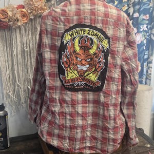 May include: A long-sleeved button-down shirt in a red and beige plaid pattern. The back features a large rectangular patch with a graphic of a horned devil figure, flames, and the text White Zombie La Sexorcisto Devil Music Vol. 1.