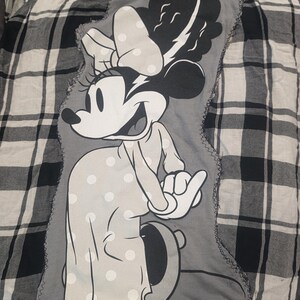 May include: A black and white plaid flannel shirt with a Minnie Mouse applique. The applique features a smiling Minnie Mouse in a polka dot dress and bow. The applique is stitched onto the shirt.