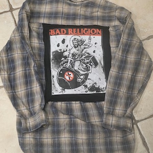 May include: Plaid button-down shirt with a large black patch featuring the text "BAD RELIGION" in red. The patch displays a graphic of a figure on a record with a symbol. The shirt has a blue, brown, and tan plaid pattern.
