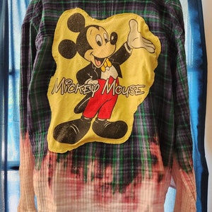 May include: A blue and green plaid flannel shirt with a yellow patch featuring Mickey Mouse. The patch has the text "Mickey Mouse" on it. The shirt has been bleached, creating a faded effect.