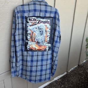 May include: A blue and gray plaid long-sleeve button-down shirt featuring a large rectangular patch on the back. The patch displays a graphic of Rob Zombie with the text "ROB ZOMBIE" and "I WAS BORN TO GO INSANE" surrounded by several skull illustrations.