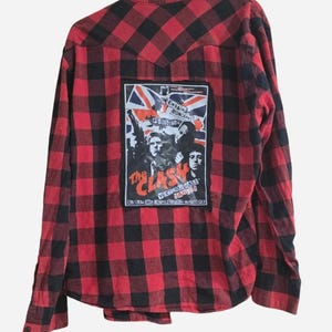 May include: Red and black plaid button-down shirt with a patch featuring the band "The Clash" and the Union Jack. The patch includes the band's name and concert details. The shirt has long sleeves and a classic collar.