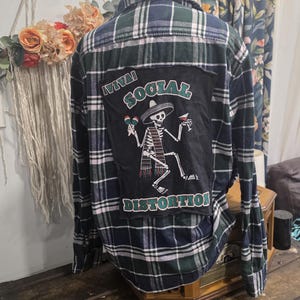 May include: A long-sleeve button-down shirt in a dark green, navy blue, and white plaid pattern. The back features a large black rectangular patch with a graphic of a skeleton wearing a sombrero, holding maracas and a drink, with the text VIVA SOCIAL DISTORTION.